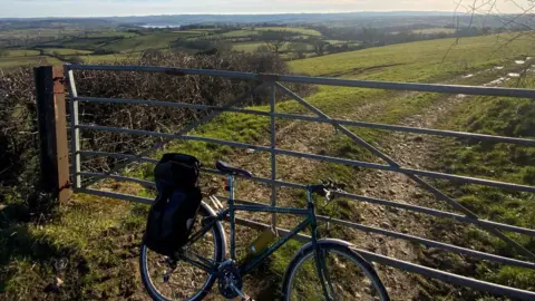 A bicycle leans against a fence at Dundry near Bristol. In the distance are the fields and woodland surrounding the lake at Chew Magna. It is a sunny day with clear blue skies