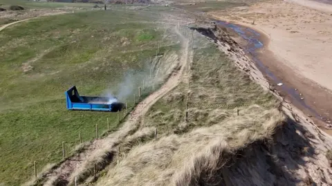 Smoke coming from small blue cordoned off structure on a grassy beach cliff. On the right hand side of the photo you can see sand. 