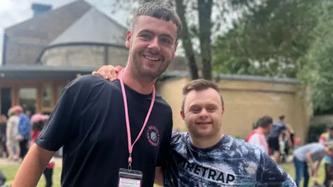 Bedford Inclusive Performing Arts Festival Volunteer Jack, smiling and looking at the camera, wearing a lanyard, and dark T-shirt, with short dark hair and stubble. He is next to James, who has his arm on his shoulder, wearing a patterned T-shirt. 