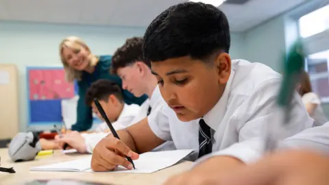 A medium shot of teenage boys in school uniforms sit side by side at their desks, working on a classroom task. The students write in their notebooks while smiling. The classroom environment is bright and typical of a modern school, with other students and teachers visible in the background. The image captures a moment of peer learning and collaboration in education. A teacher can be seen helping her students.