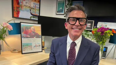 Aisha Iqbal/BBC A man wearing a blue suit and purple tie, and large black glasses, sits in front of a desk and smiles. On the desk behind him is a large picture frame depicting David Hockney and the dates 1937 to 2026. Also on the desk is a vase of flowers and a computer. There are more paintings on the wall.