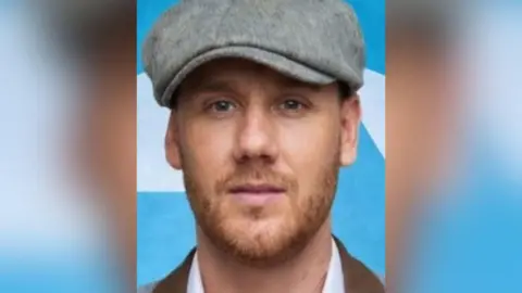 A man looking into the camera. He has a light beard and is wearing a grey flat cap. Behind him is a light blue background with white shapes on it.