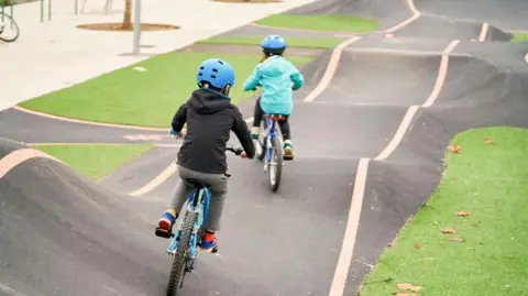 Two children ride bikes on a pump track in a park. The track has lines painted on either side and has bumps and dips. There are grass areas around the track.
