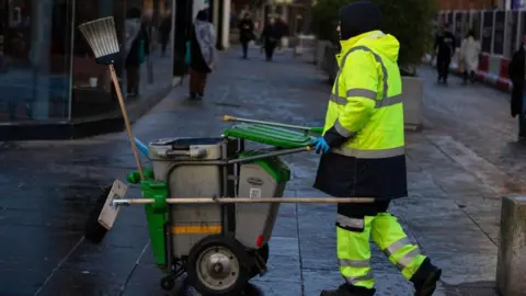 A bin worker pushes a cleaning cart through Glasgow city centre, wearing a hi-vis jacket and blue gloves. People are walking down a street behind him towards the St Enoch Centre