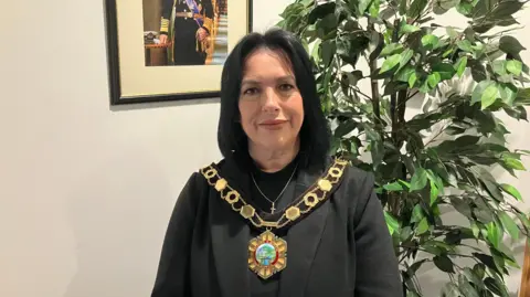 A woman with long dark hair and a dark top with a gold chain of office around her neck and a green plant behind her