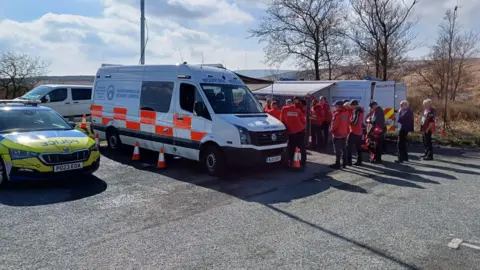 BoltonMountainRescue A police vehicle parked next to mountain rescue vehicles in a car park. A huddle of people in orange jackets can be seen gathered behind one of the vans. 