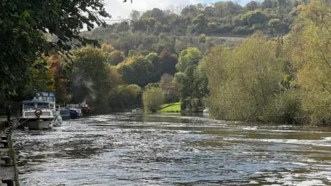 BBC A wide river with trees on both sides, and some boats moored up on the left hand side.