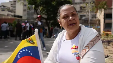 Rosa Contreras looks at the camera intently. She wears plain white clothes and holds a Venezuelan flag in her hand.