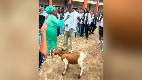 Courtesy of Promise Okoro Promise is holding a tether with the goat on the other end. His mother - in a green traditional Nigerian outfit - is gesticulating with her right hand. The are surrounded by fellow students.