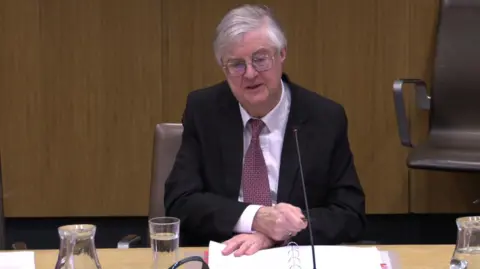 Senedd Cymru Mark Drakeford sitting at a desk in the Finance Committee, wearing glasses, a dark suit, a shirt and tie. He has a file of papers,a glass of water, a jug and a long-stemmed microphone in front of him.