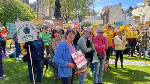 BBC/Richard Edwards A crowd gathered in a park space holding signs opposing fracking, including messages about protecting the planet and prioritising people over profit.
