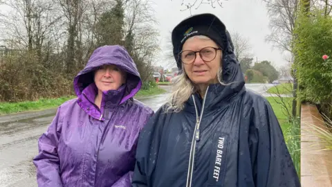 Louise Robertson and Pauline Tyler both with their hoods up.Louise is wearing a purple waterproof jacket. Pauline is wearing a dark blue waterproof, with long grey hair poking out of her hood. She is also wearing glasses.