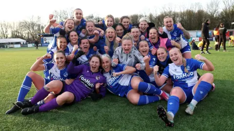 Getty Images Players of Bristol Rovers pose for a group photo on the pitch as they celebrate victory after the Adobe Women's FA Cup Third Round match against Oxford United. They are smiling and laughing and many of them are punching the air. The players are wearing the traditional Rovers kit of blue and white quarters.