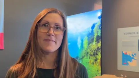 Jo Burn / BBC A woman with brown hair and glasses stood in a room with a sea bed image behind her