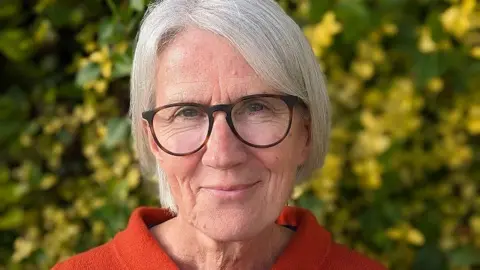 A woman with white hair wearing dark-rimmed glasses and an orange top, smiling at the camera with leaves in soft focus behind her.