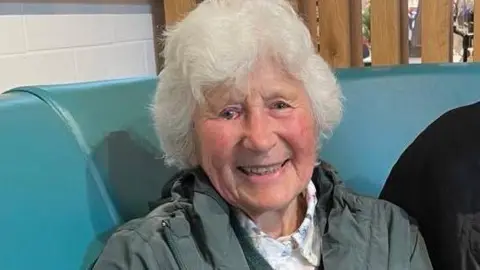 Anne Kennedy Woods, who has wavy grey hair and is wearing a green-grey coat over a white floral blouse. She is smiling broadly.