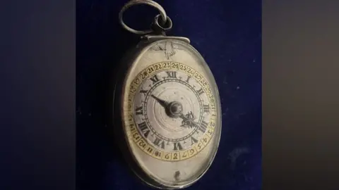 The Cromwell Museum The front of a 17th Century silver oval-shaped pocket watch, with two rows of numerals, the inner Roman numerals from I to XII, the outer Arabic numerals from 1 to 31, the watch suspended from a loop at its top, against a black background