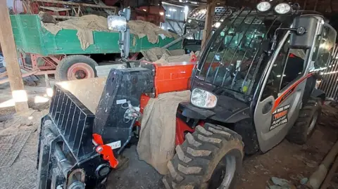 Essex Police A large plant machine, with large wheels and a glazed cabin in the middle. It is resting on an earth floor in a barn. There is a farm trailer behind it, piled with tarpaulins. 
