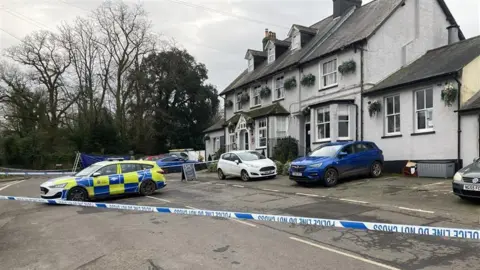 Dan MacLaren/BBC A general view of the Three Horseshoes pub in Knockholt with police car and police cordon in foreground