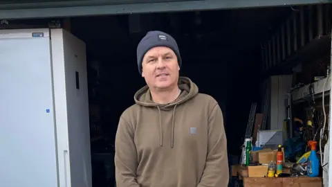 Kate Bradbrook/BBC A man smiles at the camera standing in front of an open garage. He is wearing a navy beanie hat with a brown hoodie on.