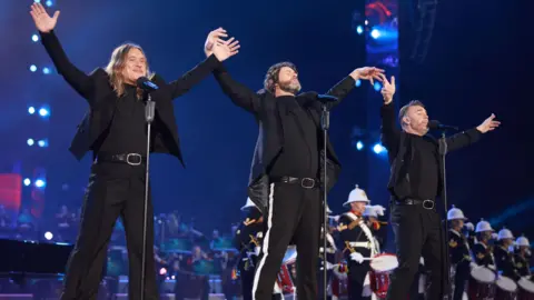 Getty Images Gary Barlow, Howard Donald and Mark Owen of Take That, all in black, perform on stage during the Coronation Concert in May 2023 in Windsor