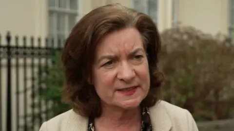 Eluned Morgan talking to someone off camera, wearing a white jacket in front of some railings and foliage which is out of focus.
