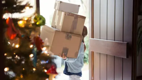 A young girl holding a pile of boxes stands by the open front door, her face hidden by the boxes. A Christmas tree is in the foreground of the image, out of focus. 