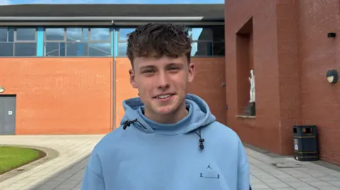 Caoimhin is smiling into the camera outside his school building which was red brick. He is wearing a blue hoody and has short brown curly hair. 