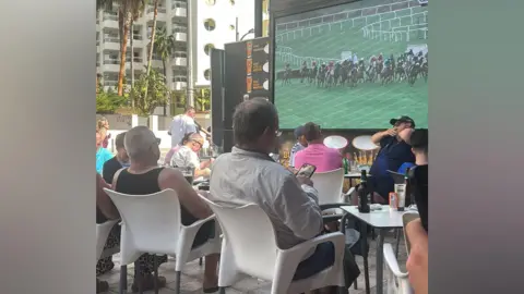 Mikey Fenton A group of people watching a horse racing event on a large TV screen outside a pub.