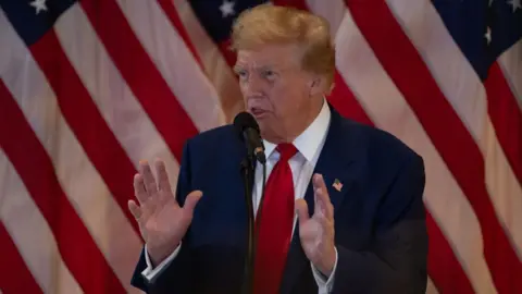 Trump speaking to reporters and gesturing with his hands in front of a flag