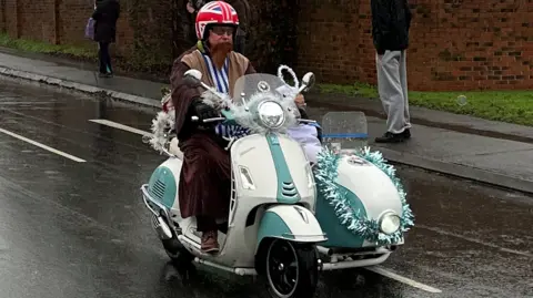A man riding a green and white scooter wearing a fake beard, gown and union flag crash helmet taking part of the Toy Run.