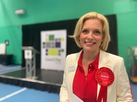 The Labour MP for South Derbyshire Samantha Niblett smiles on the night she is elected, standing in a sports hall wearing a cream blazer and a red Labour Party rosette 