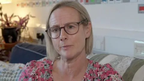 Heather Richards has short blonde hair and round glasses. She is wearing a pink floral top.