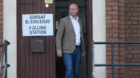 Dan Thomas stepping out of a door in jeans, a jacket and a shirt, with a sign that reads polling station next to him.