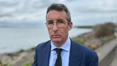 Andrew Muir, a man with grey hair and glasses, wearing a dark grey suit, white shirt and black tie.