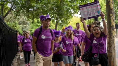 Kidney Research UK A procession of people wearing purple Kidney Research UK t-shirts walking alone a pavement. One is holding a white placard with the words "WALKING for my MARIA" written on it.