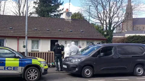 Uniformed police officers and forensic investigators wearing white protective suits stand outside a house in Stream Street, Downpatrick.  The house is a semi-detached or end-of-terrace bungalow.  There are trees behind the houses and church with a tall spire in the distance.  A liveried blue and yellow police car is parked outside the house and other vehicles are parked in the street. 