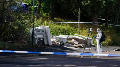 PA Media A man in a white forensic suit stood in front of a tripod and camera. He is at the scene of a woodland area. There are various bits of old furniture scattered. The area is cordoned off with police tape. 