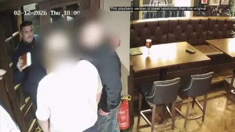 Three men are standing inside of a pub at the entrance which is on the left. Two people are blurred and the other is holding a Bible and facing the CCTV. 