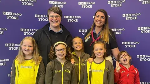 Two women stand behind four girls wearing Brownies uniform in front of a BBC Radio Stoke background 