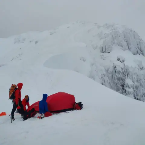 Glencoe MRT Rescuers stand next to a red bivvy on a snowy mountain top.
