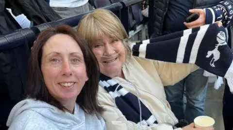 Two women sat in a football stadium wearing football scarves.