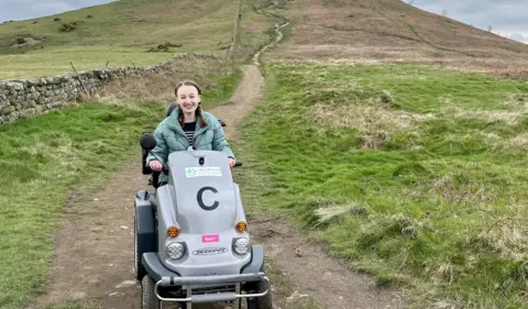 Pippa Stacey A young woman with short brown hair tied in two plaits sits on a large grey off-road mobility scooter. She wears a green puffed jacket and a blue and white striped jumper. She is surrounded by countryside scenes - an unpaved path, leading up a grassy hill. 