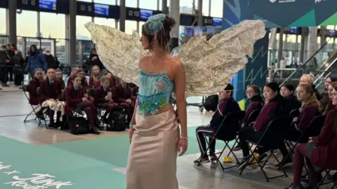 A student walking the catwalk in a light colour skirt and blue corset made from recycled material. She also has wings made of old plastic. 