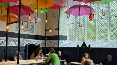 Peterborough Cathedral People sitting on wooden benches inside the cafe, lined with colourful umbrellas on the ceiling.