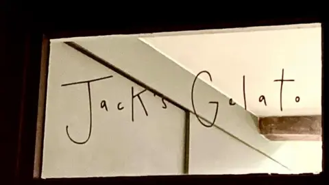 Brian Farmer/BBC Sign on a fanlight at a Jack's Gelato ice cream shop in Cambridge. The words "Jack's Gelato" are written in black handwriting on a window. Part of a wall and ceiling, and a beam, are visible through the glass. 