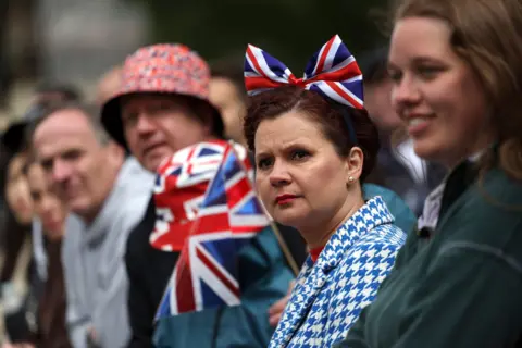 EPA A person looks on while wearing a blue and white checkered suit and a union flag bow on her head, with other crowd members waving British flags alongside the side of the Mall during the procession.