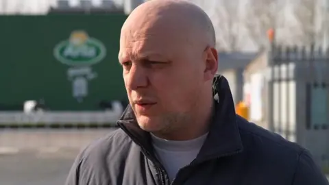 A man in a dark coat stands outside the green Arla factory building. He is bald and looking away from the camera.