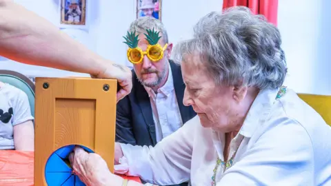 PA Media Taskmaster host Alex Horne looks on as resident Diana takes part in a test at Seccombe Court care home in Adderbury.