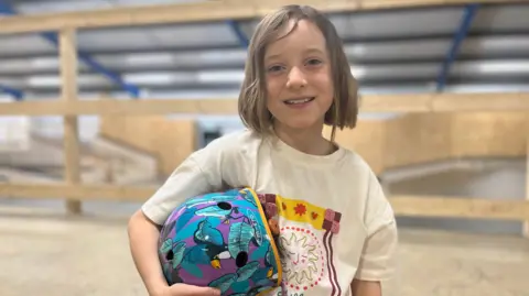 Aimee Dexter/BBC Grace is sitting on a ramp. She has short blonde hair and is wearing a white T-shirt which has a pattern it. She is also holding a purple and blue helmet, which has leaves and a bird on it.